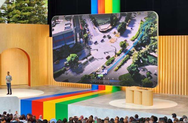 Photo of the stage at Google I/O keynote. Alphabet CEO Sundar Pichai is standing off to the left. A large screen shows a new feature in Google Maps, as 3D render of the route that the app is suggesting to a user. It shows a visualization of a city environment with a blue line and an arrow along a sidewalk. It shows the level of traffic, the temperature and the time of day.