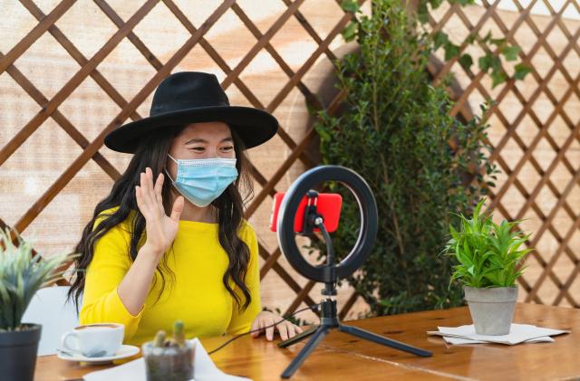 Happy Asian woman wearing face mask and vlogging online using smartphone cam and influencer led while drinking a coffee sitting in bar during corona virus outbreak