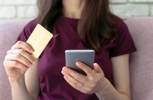 Close up cropped photo of adult and confident woman sitting in cafe with bright light and modern interior. Female using plastic card and holding smartphone in hands