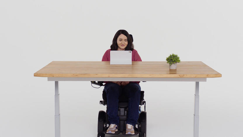 A person in a wheelchair sitting in front of a table where an iPad is set up. They are facing the iPad and smiling.