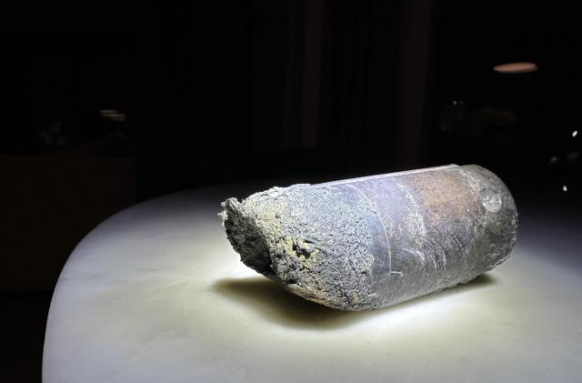 Photo of a stanchion from an ISS space dump. The object, a gray-ish cylinder with crumbled / burned edges, sits on a white table with black background.