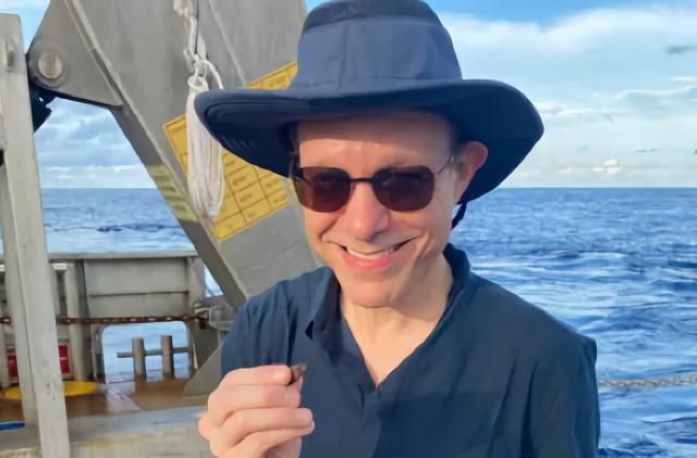 Photo of Harvard astrophysicist, on a boat with sunglasses and hat, holding a small object in his hand.