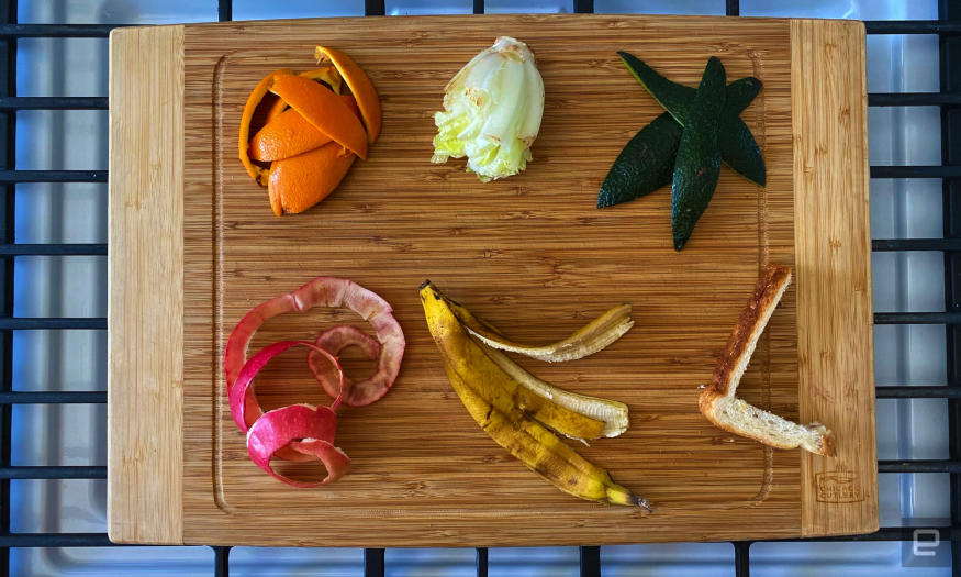 Food scraps, including lettuce, sandwich crust and peels from an orange, apple and banana, are arranged on a bamboo cutting board. 