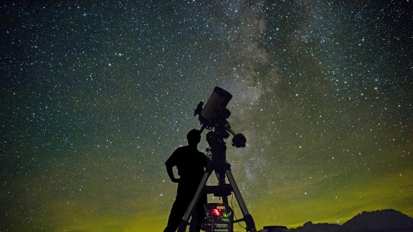 An Astronomer with a telescope stands in silhouette against a starry night sky with a greenish yellow glow at the horizon line.