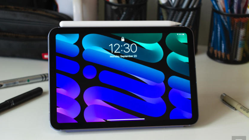 Photo of the 6th-generation iPad mini sitting on a desk with its Apple Pencil magnetically attached to its side. Its colorful blue / green lock screen (with cursive "mini" on the screen) is displayed. Pens all around.