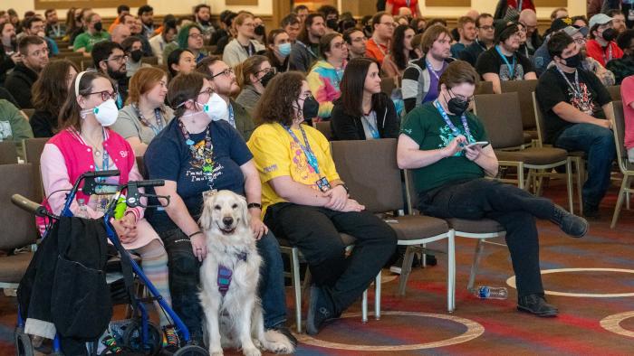 A room full of audience members at a Games Done Quick charity speed-running event. In the front row, someone holds a cream-colored golden retriever, who smiles for the camera.