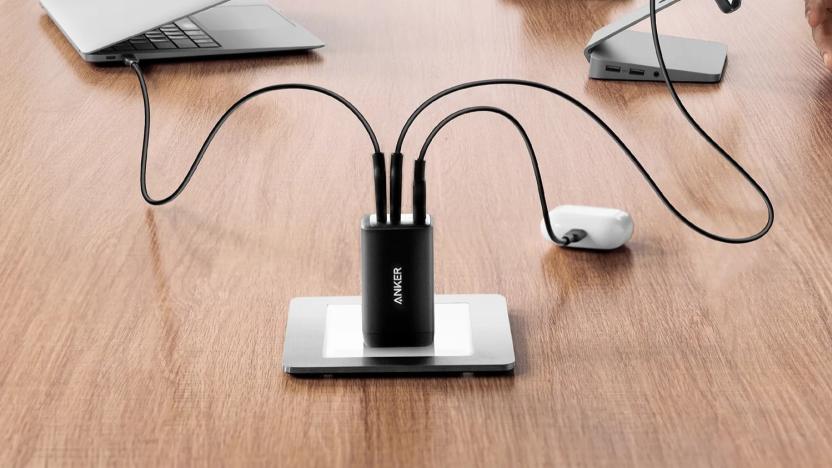 A small black wall charger, the Anker 735, shown plugged into an outlet on a brown wooden desk, with cables attached charging a laptop, smartphone and pair of wireless earbuds.