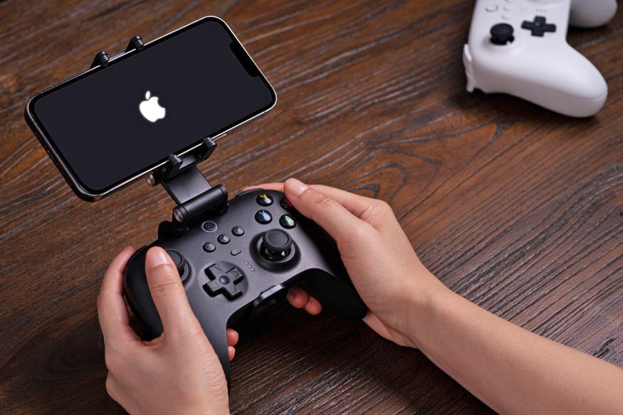 Two hands hold an 8BitDo controller that's paired with an iPhone showing the Apple logo on the screen.