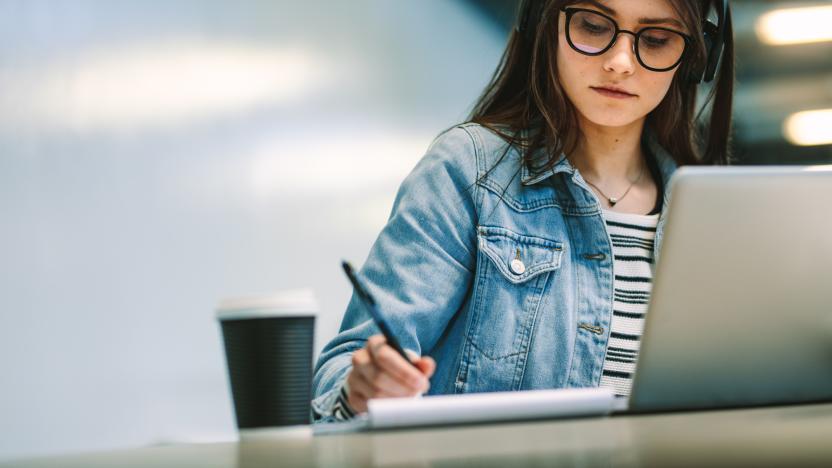 University student writing in a book while sitting at desk with laptop and coffee up at college campus. Female student studying at college library.