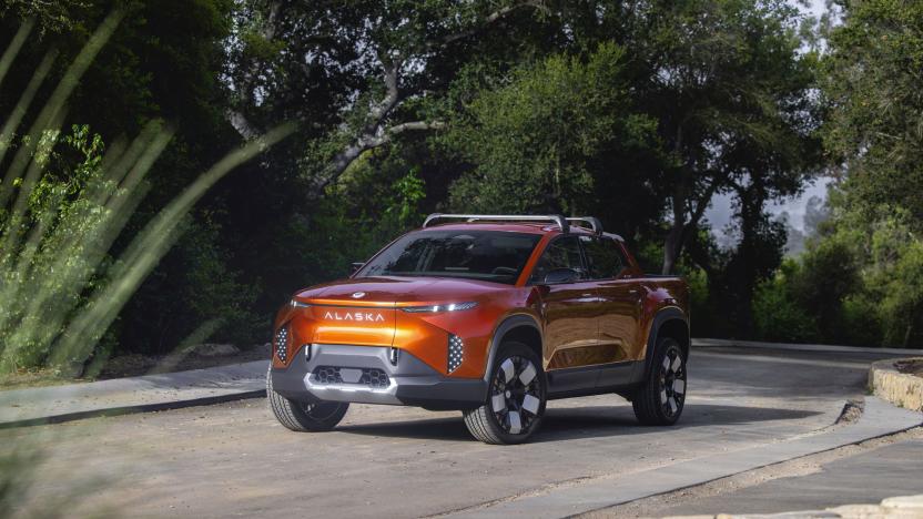 Image of an orange Fisker Alaska flatbed EV driving down a quiet road in front of some dark green foliage.