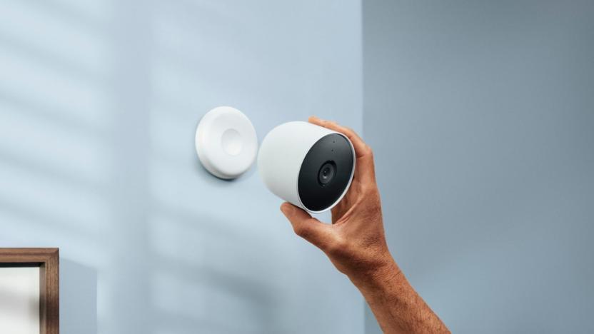Product lifestyle marketing image of the Google Nest Cam Indoor / Outdoor. A hand reaches up to mount the camera magnetically to a wall mount.