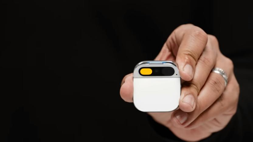 The hand of co-founder Imran Chaudhri holding the Humane AI Pin. Closeup. The small device has a white face, and is square with round corners and a section at the top for a camera.