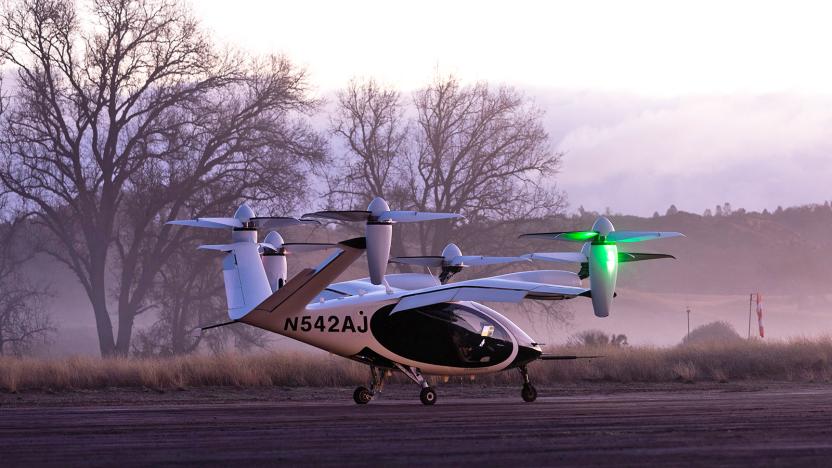 The Joby Aviation electric air taxi sits on a runway during pinkish pre-dawn hours with mist hanging among the trees in the background.