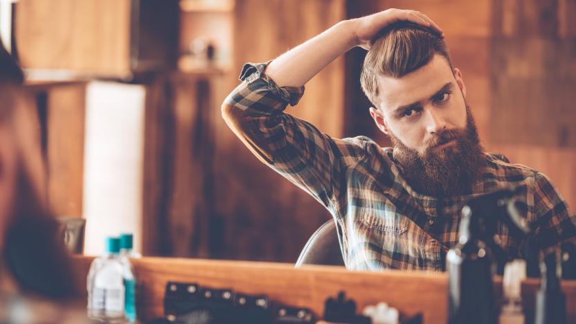 Handsome young bearded man looking at his reflection in the mirror and keeping hand in hair while sitting in chair at barbershop