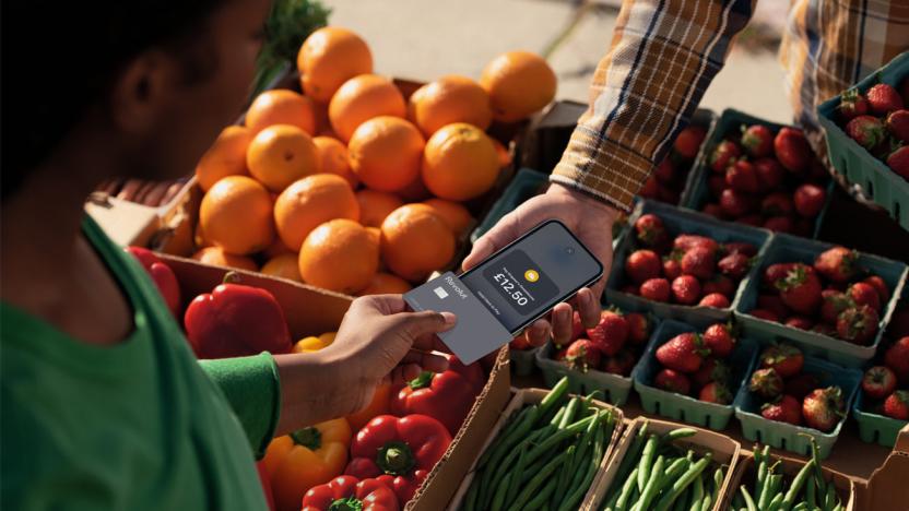 A person wearing a green shirt holding a credit card over an iPhone to pay for their purchase. Oranges, bell peppers, strawberries and string beans in containers are in the  background.