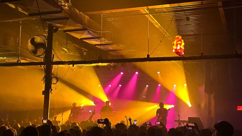 A band called Thrice is seen playing on stage from the perspective of an audience member. People are holding up phones to film them and the band is lit by pink and yellow spotlights.