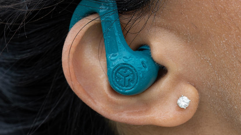 A green-blue earbud with an earhook and water on it is positioned inside a person's ear.