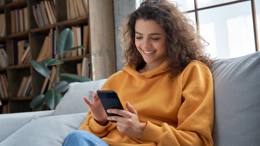 Happy millennial hispanic teen girl checking social media holding smartphone at home. Smiling young latin woman using mobile phone app playing game, shopping online, ordering delivery relax on sofa.