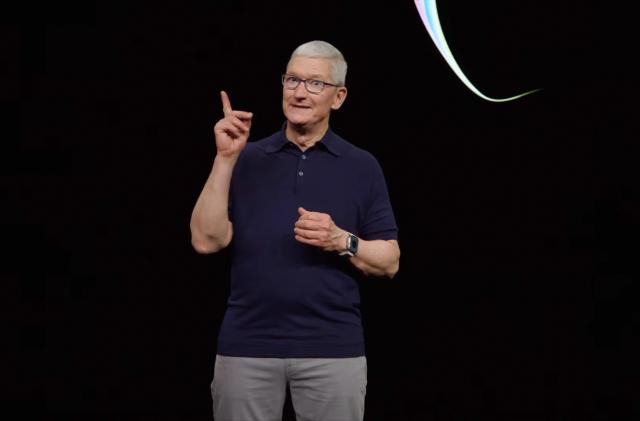 Tim Cook standing in front of a black backdrop (with segment of an Apple logo visible) holding up his index finger