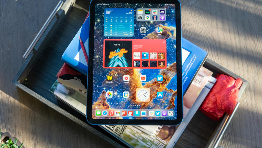 The 10th-generation iPad rests outside on top of a stack of books, on a wooden desktop, with its screen active, displaying a colorful home screen with various iPadOS widgets and apps.