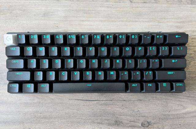 The Logitech G Pro X 60 wireless gaming keyboard in black sits on a wooden tabletop with light blue RGB backlighting displayed through its keycaps.