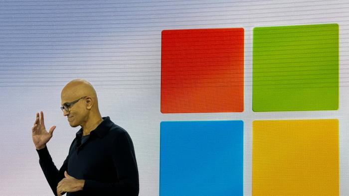 Microsoft CEO Satya Nadella onstage in front of a Windows logo. He stands to the left of it, gesturing passionately.