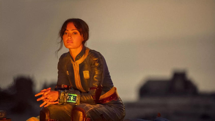 Ella Purnell (Lucy) in a still from the Fallout TV series. She sits, reflecting in front of a blurred desert landscape.