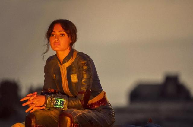 Ella Purnell (Lucy) in a still from the Fallout TV series. She sits, reflecting in front of a blurred desert landscape.