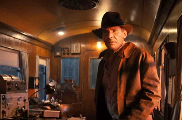 Harrison Ford looks at something (or someone) off camera as he stands in what appears to be an old train car with communications gear off to the side. It's dark out and there's soft yellow lighting inside.