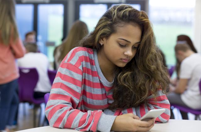 Female student sitting on her own at school. She has a smartphone in her hand and a stressed expression on her face.