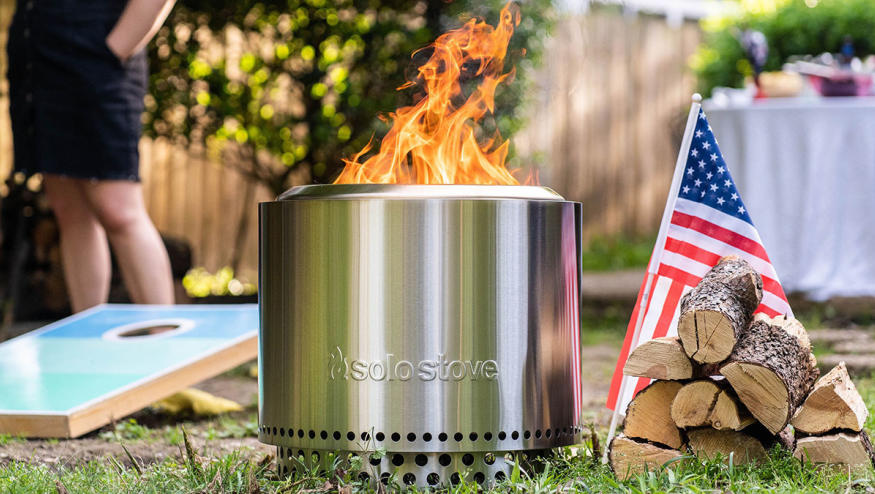 A Solo Stove fire pit sitting on grass next to fire wood and an American flag at a backyard party.