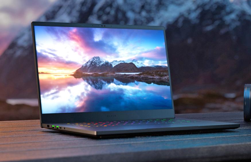 The Razer Blade 15 laptop with 240Hz OLED display seen on a picnic tabletop with mountains in the background and an onscreen image of a larger mountain view with dramatic sky and a reflective lake surface in the foreground.