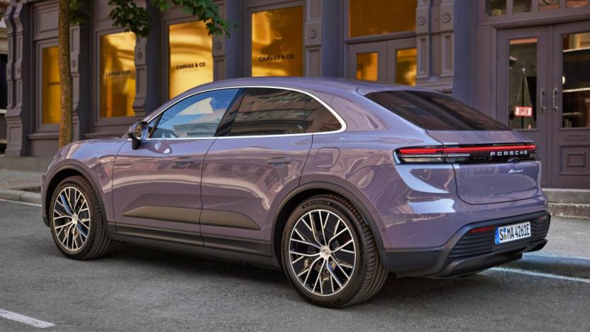 A purplish-grey Porsche’s all-electric Macan SUV is parked on the side of a road in front of doorways and windows.