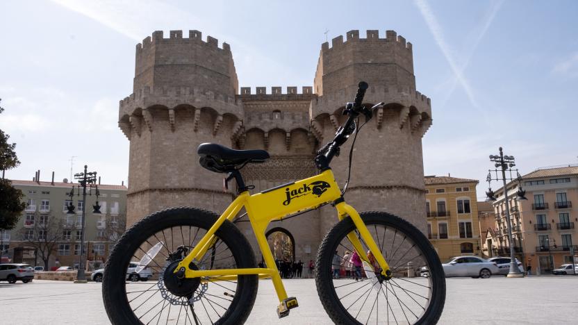 The JackRabbit ebike in yellow on its own in an empty plaza ini front of a small castle-like structure with two turrets.