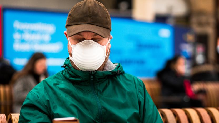 Close up color image depicting a young caucasian man in his 30s wearing a white protective face mask - to protect himself from flu viruses and the coronavirus - sitting and waiting on a bench at a railroad station in the city of London. He is wearing casual clothing, a green rainmack and messenger bag. In the background other people are defocused.