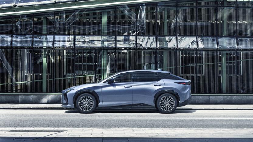 The Lexus RZ sport sedan in light metallic blue parked in front of a modern office building that's mostly made of glass windows. 