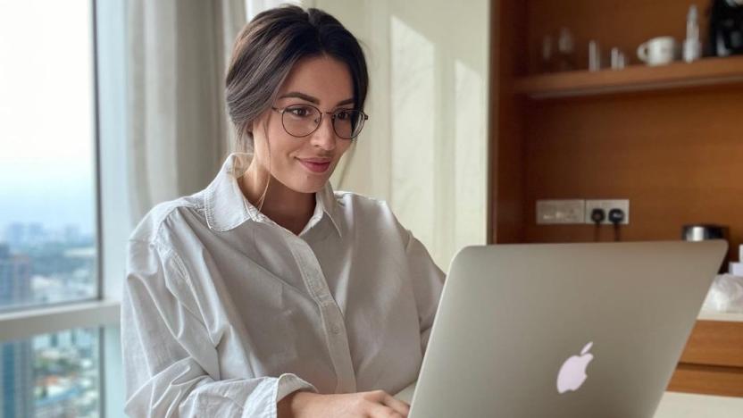 A woman sits at a table on her silver MacBook. 