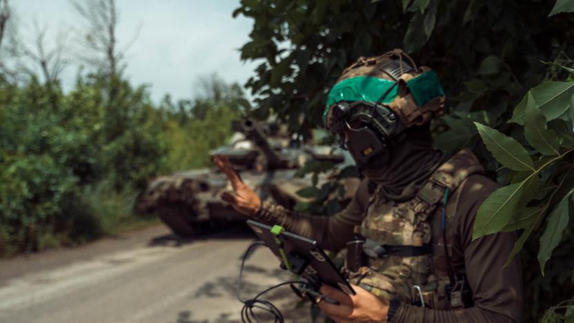 A Ukrainian soldier operating a drone. Forest, next to a street. The solider is holding a tablet with wires as a friendly tank sits behind.