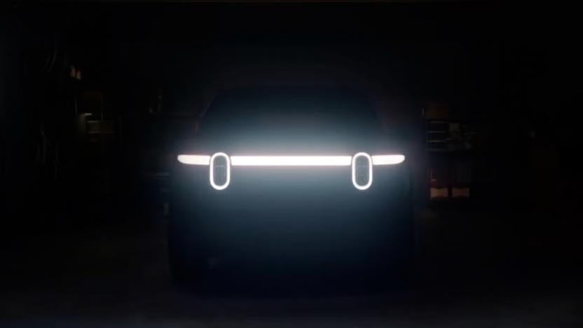 Teaser image for the Rivian R2 EV. Two vertically oval headlights along a sleek horizontal light bar hint at the front of the vehicle. Otherwise nearly black image of a garage.