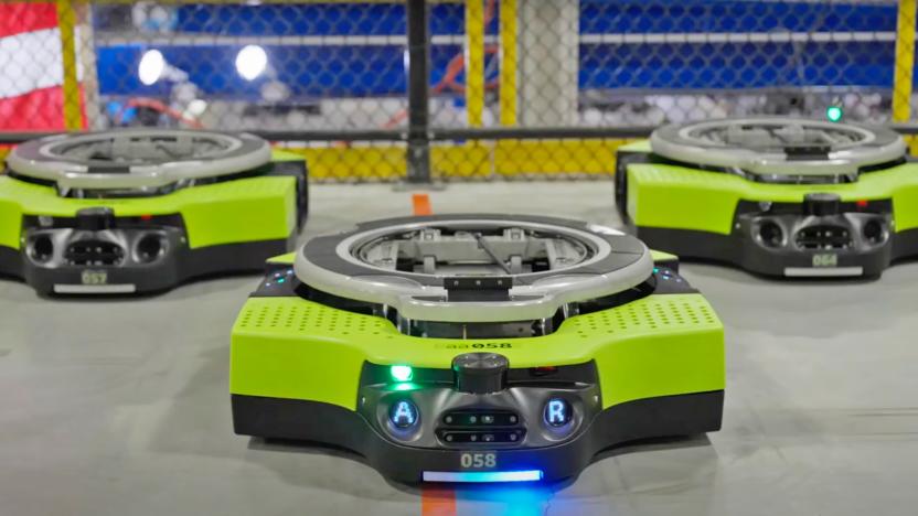 A trio of Amazon's fully autonomous Proteus warehouse robots face forward towards the camera as they sit on the floor, looking like a yellow Roomba vacuum.
