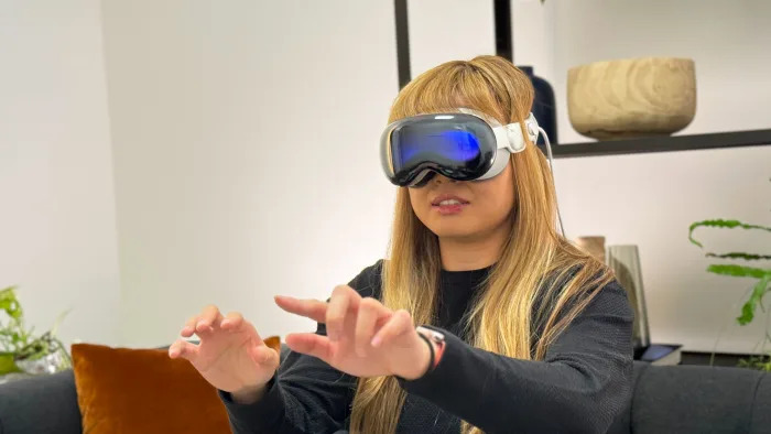 A woman with blonde hair with the Apple Vision Pro on her face, her hands are outstretched with her fingers held in a typing position.