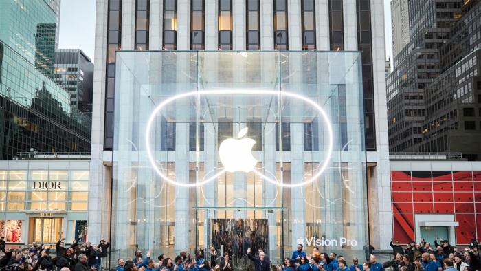 The facade of an Apple Store with the Apple logo lit up and surrounded by the shape of the Vision Pro headset.