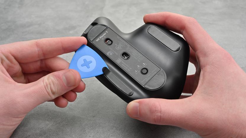 Photo of a person holding the Logitech MX Master 3 mouse with a blue spudger in the other hand pulling open a tab to open the device.