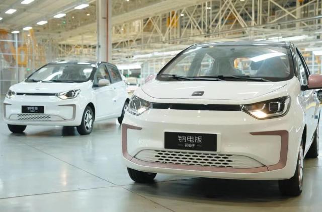 Large car factory or showroom floor featuring two small white electric vehicles. Scaffolding and other cars visible in the background.