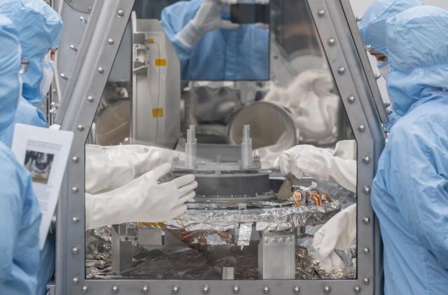 Scientists in blue overalls standing around a glass container with their gloved hands inside.