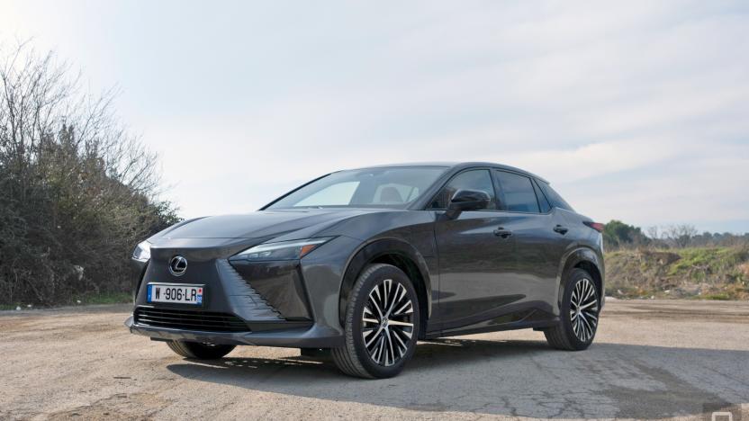 A Lexus RZ 450e in grey is parked at an angle in the middle of a dirt road with sparse grass and bushes flanking it on either side.