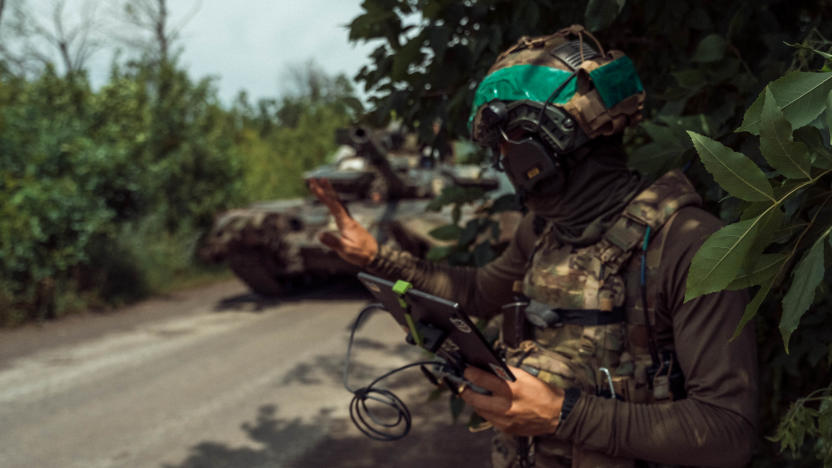 A Ukrainian soldier operating a drone. Forest, next to a street. The solider is holding a tablet with wires as a friendly tank sits behind.