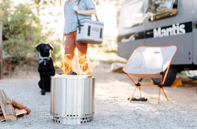 The Solo Stove Ranger fire pit sits on the ground outside with a fire lit inside of it. There is a camper, a camp chair, a black dog and a person carrying a cooler in the background. 