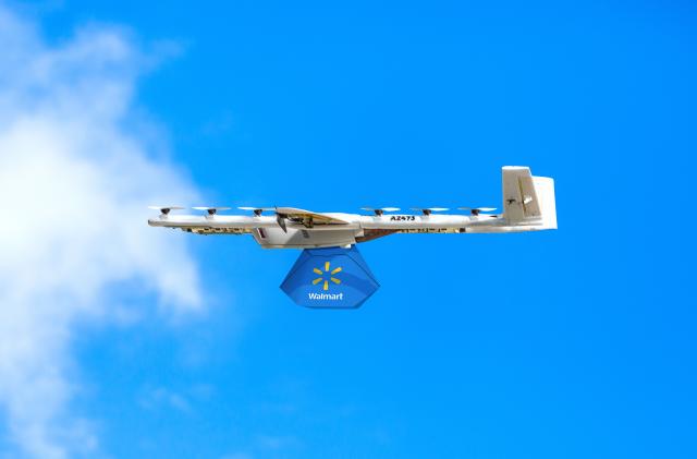 A white delivery drone carrying a blue Walmart bag soaring across a blue sky. Side view of the drone as it flies right to left across the frame.