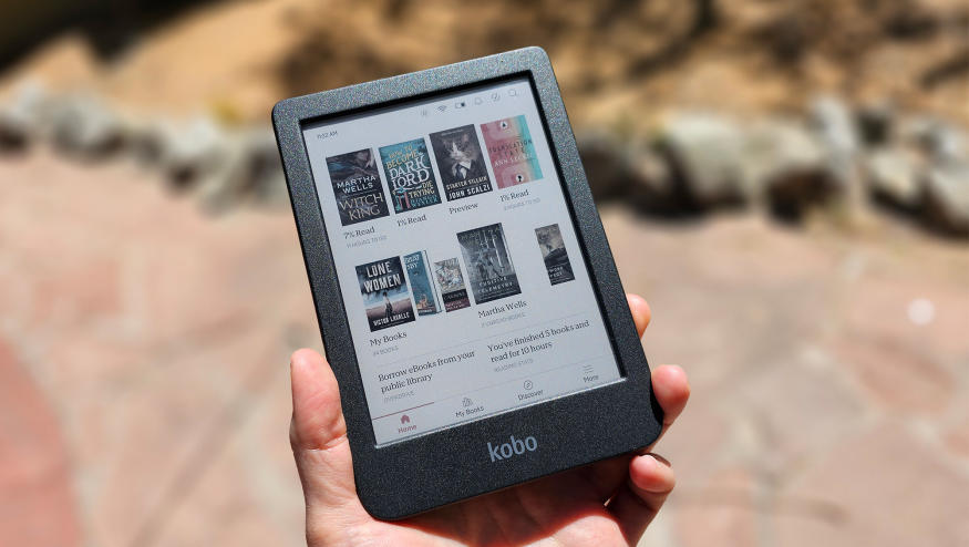 A person holds the Kobo Clara Colour in the sun. The color covers of some books are displayed on the screen. There are rocks in the background. 
